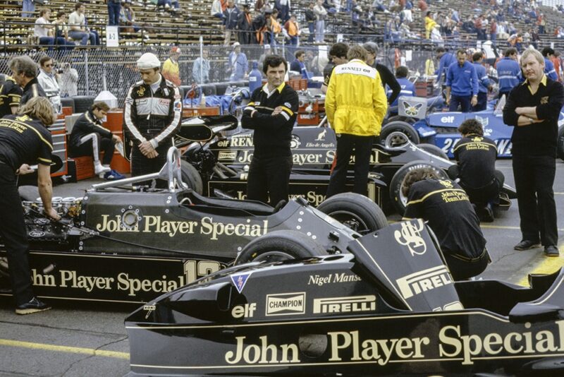 Nigel Mansell, half-wearing a balaclava, looks over at mechanics working on his Lotus 92 Ford.