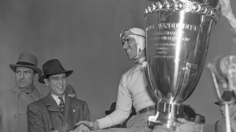 George Vanderbilt, donor of the cup, shakes hands with Tazio Nuvolari, of Italy, after the latter had won the George Vanderbilt trophy, (the huge cup shown), and first prize money, in the first Columbus Day race at the Roosevelt Raceway, Westbury, L.I., Oct. 12. Nuvolari beat out a field of international racers to win the grueling and dangerous race.