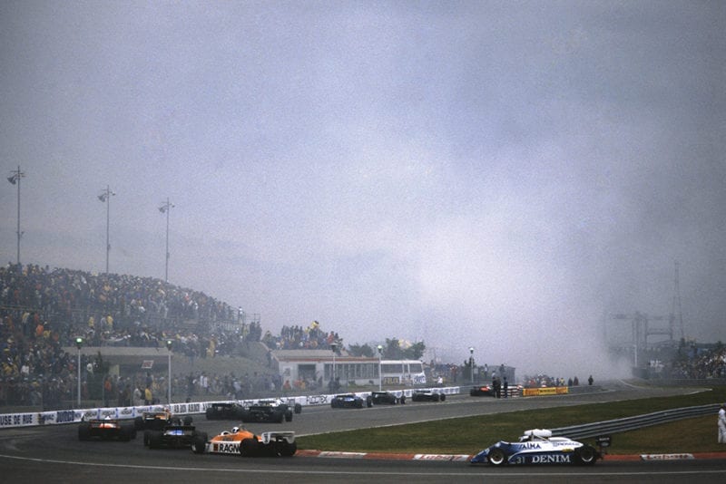 Cars return to the pits as smoke rises from the startline accident in which Riccardo Paletti lost his life.