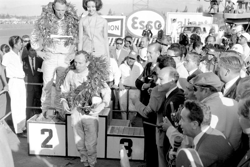 Race winner Dan Gurney stands on the podium with 2nd placed John Surtees standing below. Stirling Moss stands to the right with the media.