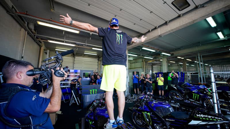 Fabio Quartararo stands on his Yamaha with arms outstretched in his pit garage with crew