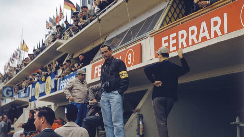 Phil Hill standing with his camera around his neck in the Ferrari pits