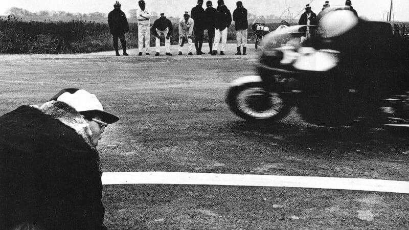 Soichiro Honda kneels at corner to watch his motorcycle passing