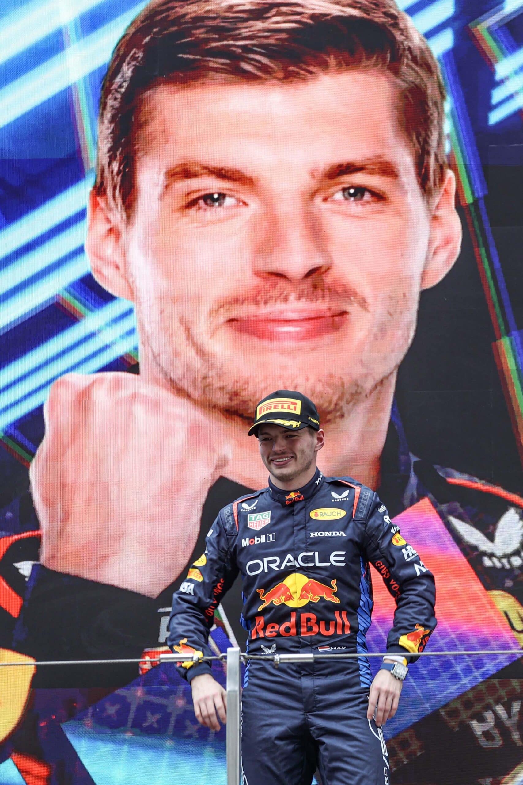 Max-Verstappen-smiles-on-the-2024-F1-Chinese-Grand-Prix-podium-in-front-of-a-ginat-screen-showing-his-face