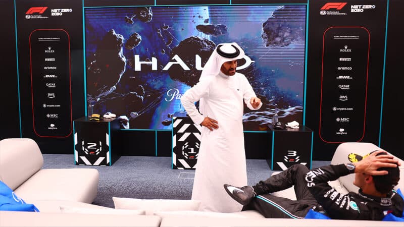 A standing Mohammed Ben Sulayem points at George Russell who is reclined on a sofa